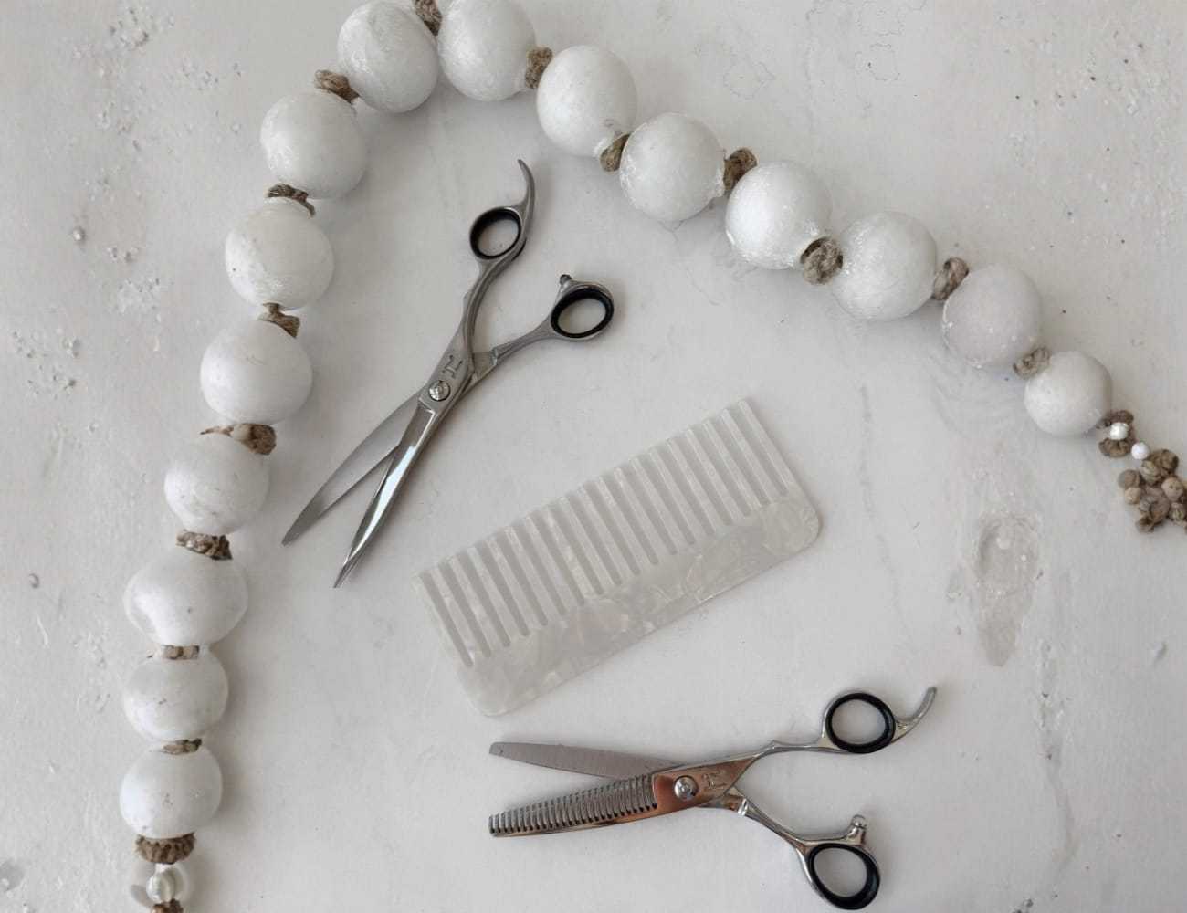 Hairdressing scissors and comb beside a string of white decorative beads on a textured surface.